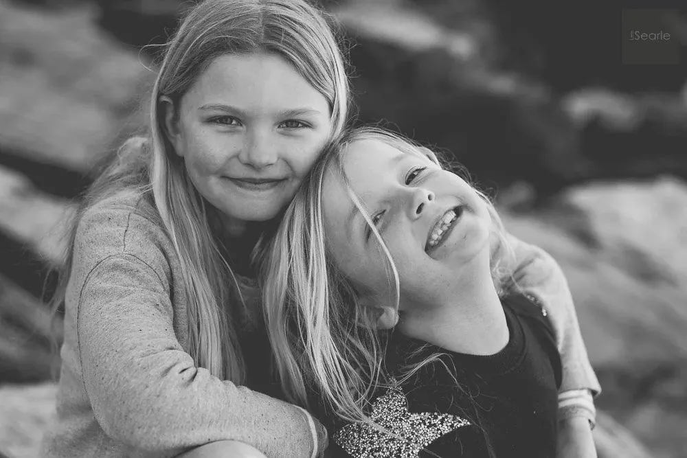 Black and white portrait of two sisters leaning together and laughing on the Cornish coast