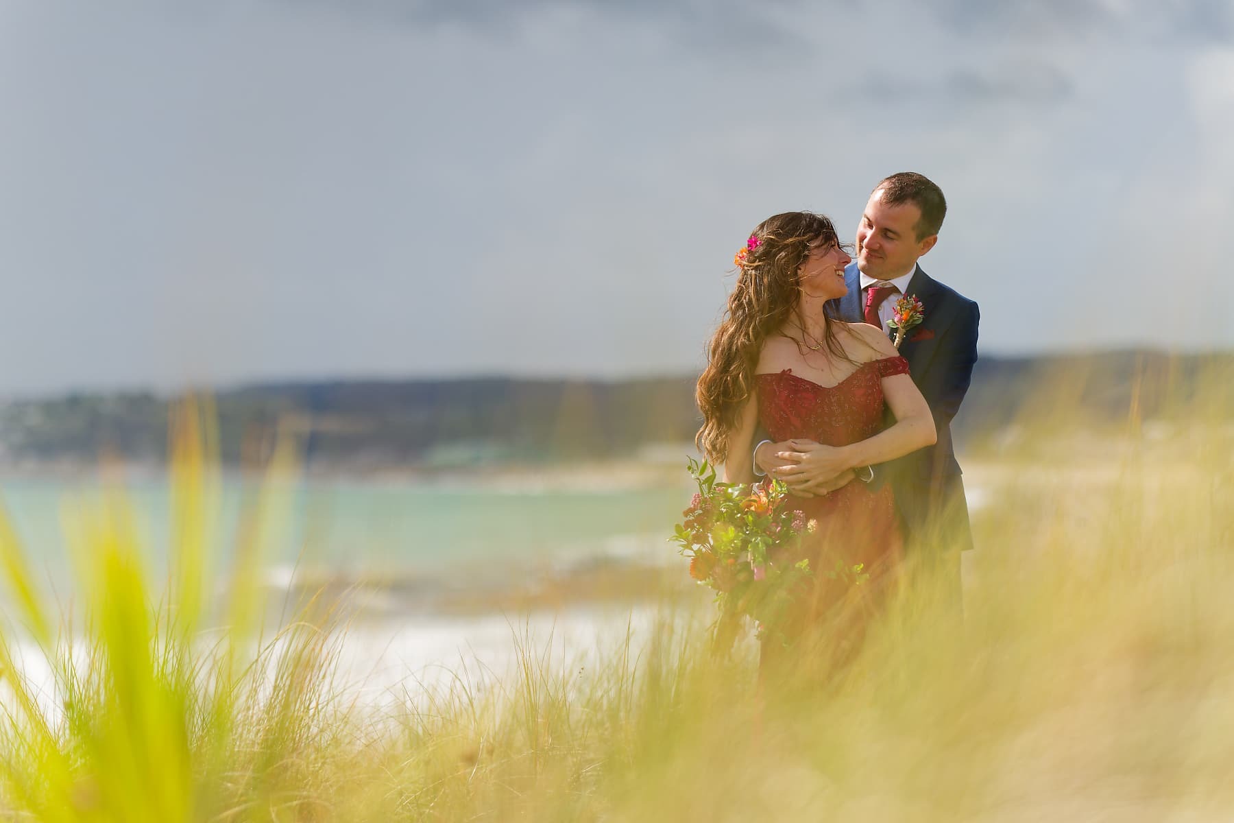 Bride and groom embracing among coastal grasses near Penzance after their wedding