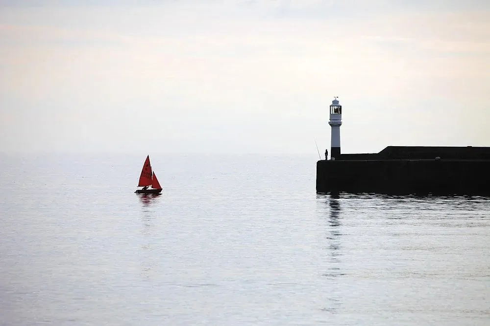 Red-sailed boat passing a harbour lighthouse on a still, misty morning in Cornwall